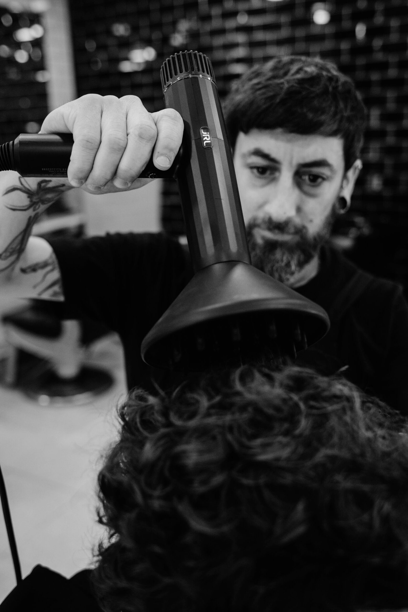 Fotografía en blanco y negro de un barbero usando un secador de pelo en los rizos de un cliente con bigote enrollado; ambos están concentrados en el proceso dentro de una barbería.