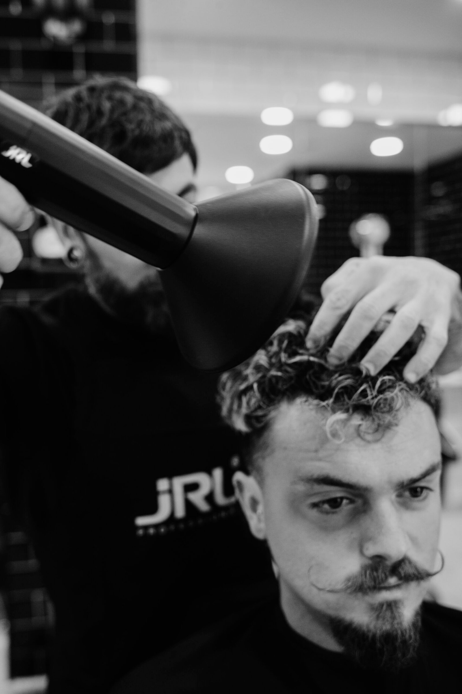 Fotografía en blanco y negro de un barbero usando un secador de pelo en los rizos de un cliente con bigote enrollado; ambos están concentrados en el proceso dentro de una barbería.