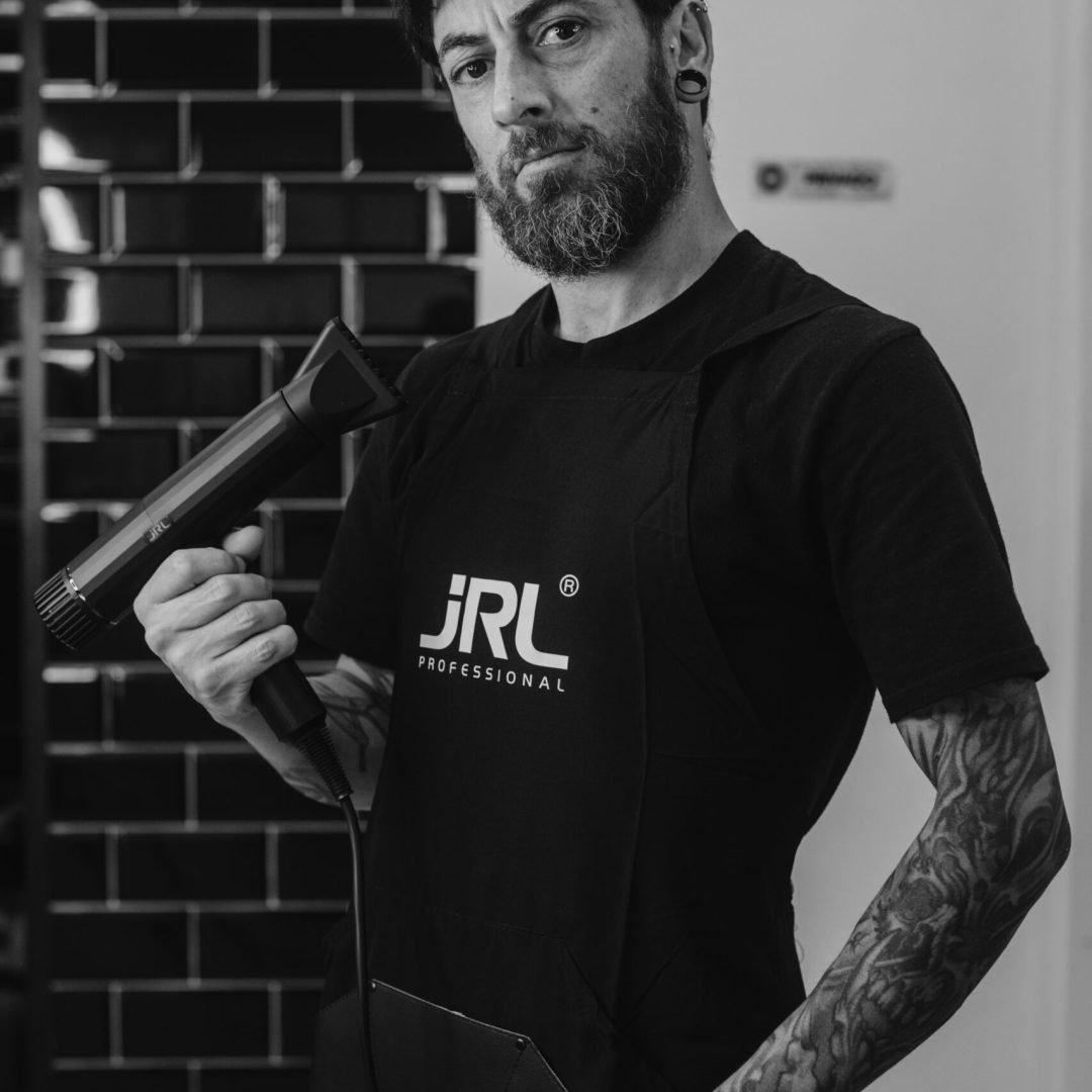 Fotografía en blanco y negro del mismo barbero tatuado de imágenes anteriores, sosteniendo una herramienta de peluquería y vistiendo delantal con el logo de “JRL”.